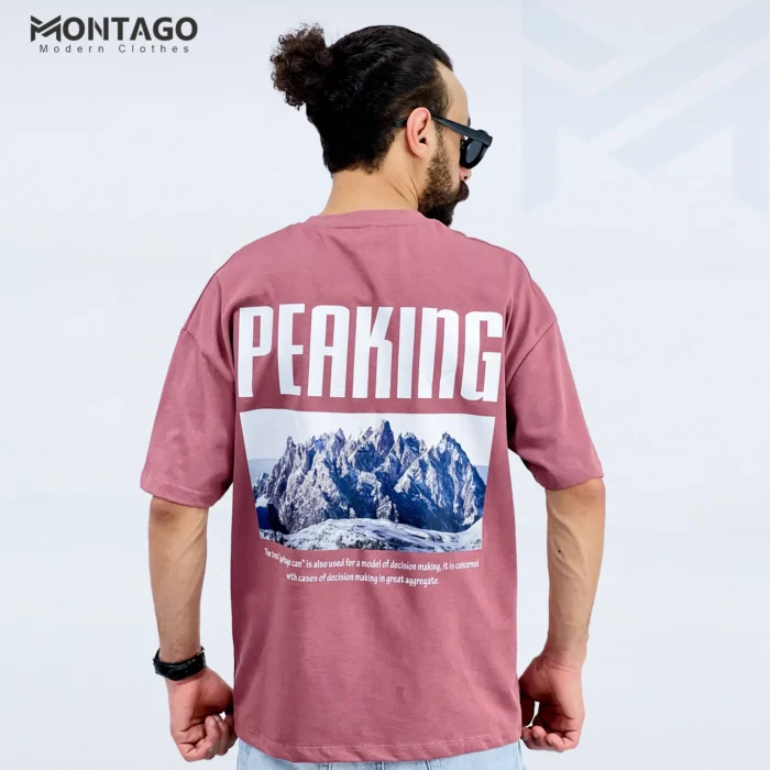 شاب يرتدي تيشيرت وردي من الخلف بطبعة كلمة PEAKING وصورة جبال، من تصميم Montago.